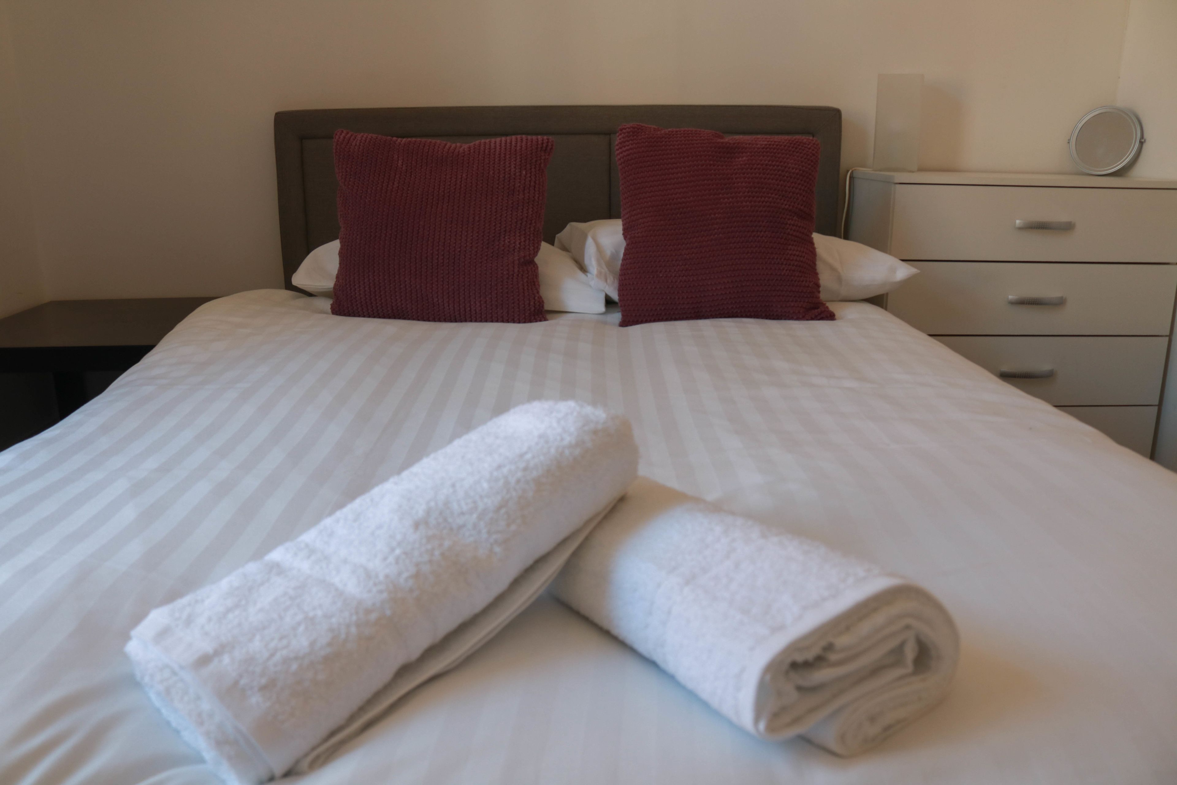 Una cama bien hecha con sábanas blancas y dos almohadas rojas, una mesita de noche con una lámpara y un pequeño mueble con cajones y un espejo.