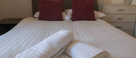 Una cama bien hecha con sábanas blancas y dos almohadas rojas, una mesita de noche con una lámpara y un pequeño mueble con cajones y un espejo.
