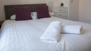 Un letto rifatto con cura, lenzuola bianche e due cuscini viola, un asciugamano bianco e un comodino con una lampada e una piccola sveglia.