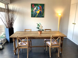 Uma área de refeições com mesa de madeira e quatro cadeiras, uma planta em vaso e um quadro colorido na parede.