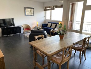 Uma sala de estar moderna com uma mesa de jantar de madeira, assentos acolchoados, uma televisão e uma grande janela que proporciona luz natural.