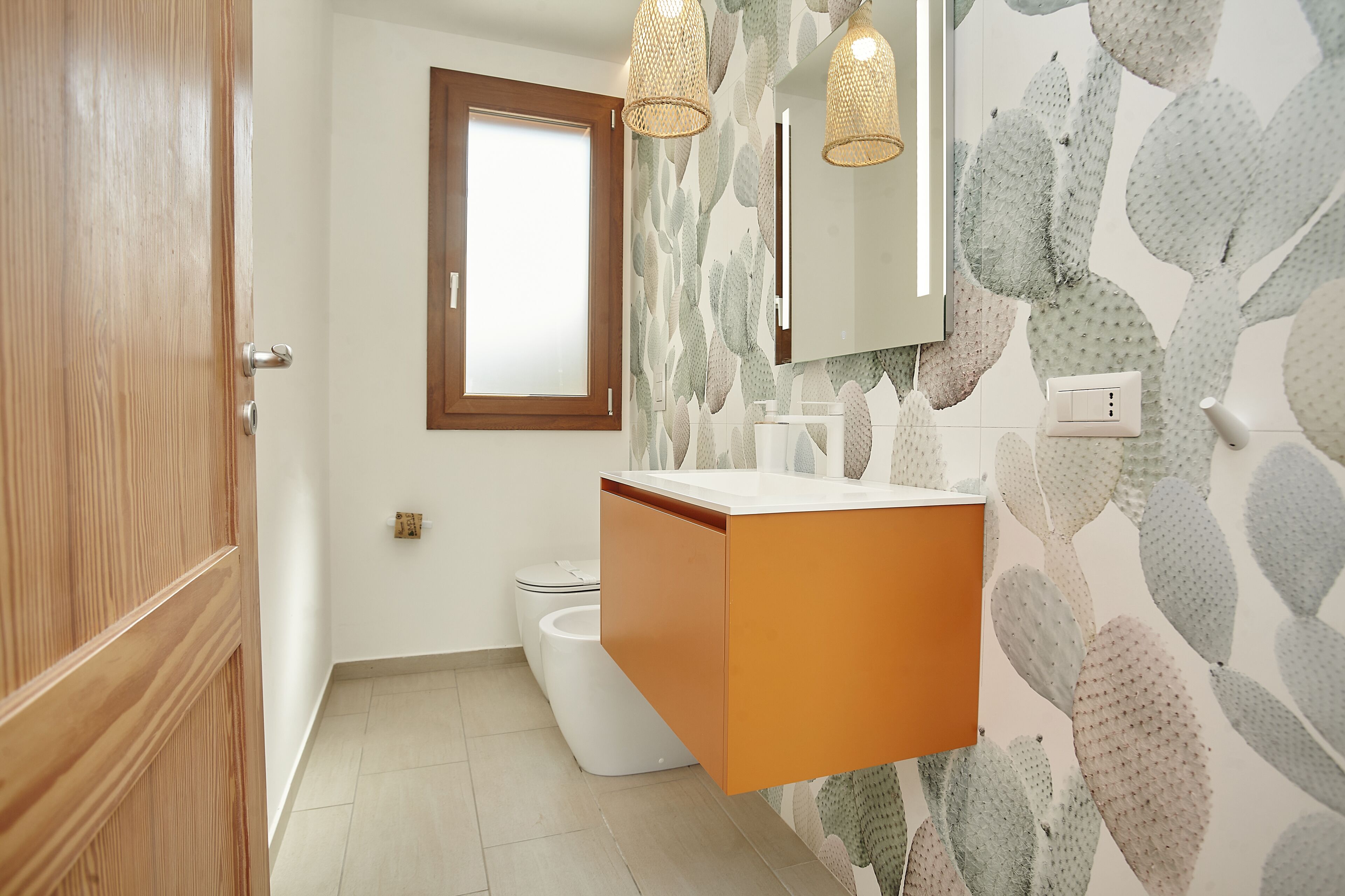 A bathroom with a wooden door, a white toilet, a wall-mounted sink with a wooden vanity, and a cactus-patterned wall.