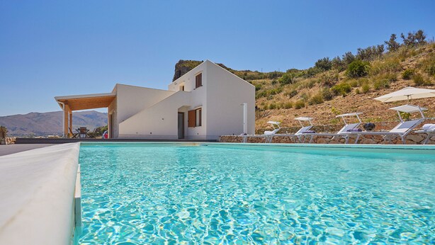 Un área moderna junto a la piscina con una pileta cristalina, un edificio blanco y vistas a una colina.