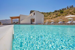 A modern poolside area with a clear pool, a white building, and a hillside view.