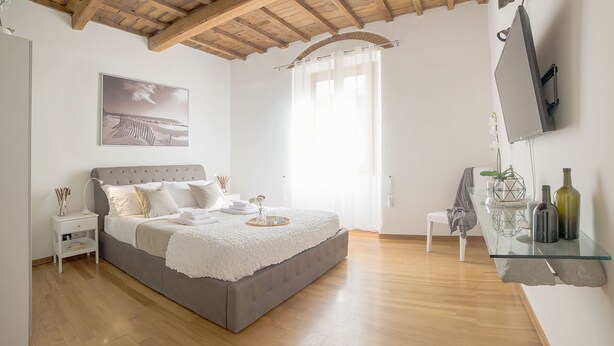 Een slaapkamer met een groot bed, een nachtkastje, een aan de muur bevestigde televisie en een glazen plank met flessen.