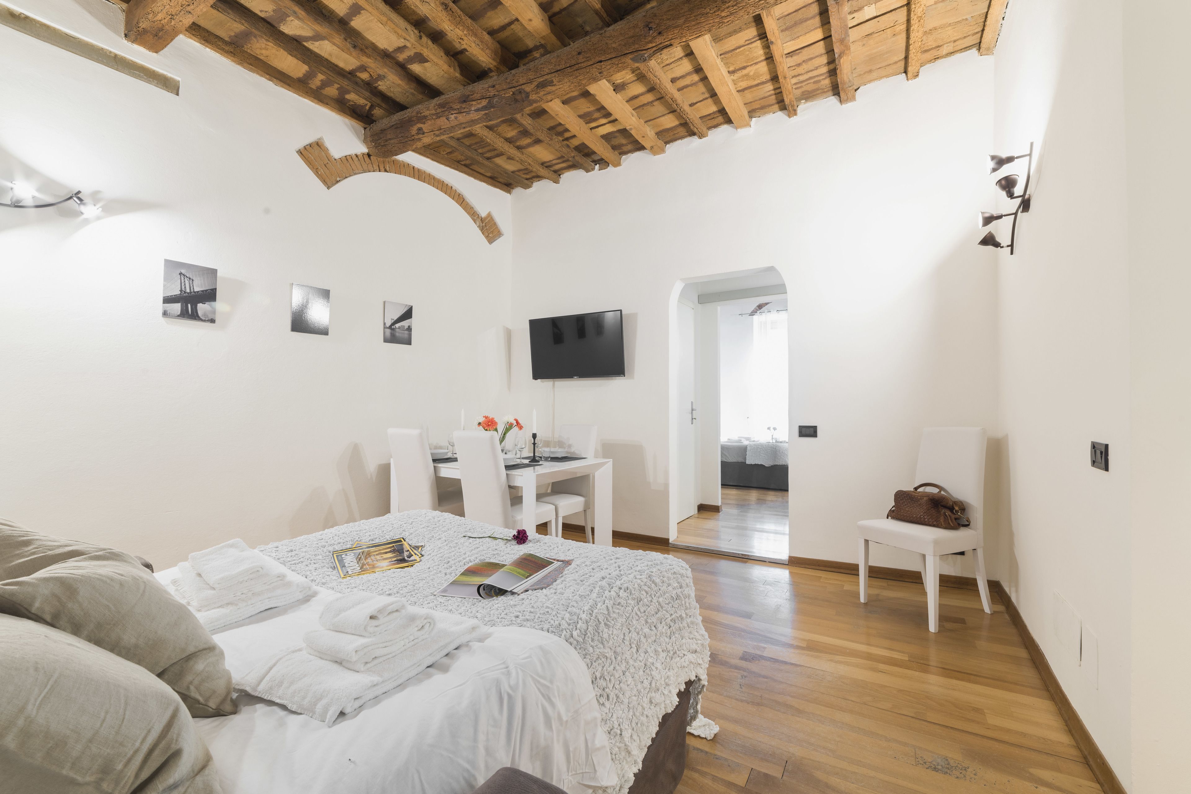 Ein Schlafzimmer mit Holzboden, einem Bett mit weißer Bettwäsche, einem Schreibtisch mit Stühlen und einem Fernseher an der Wand.