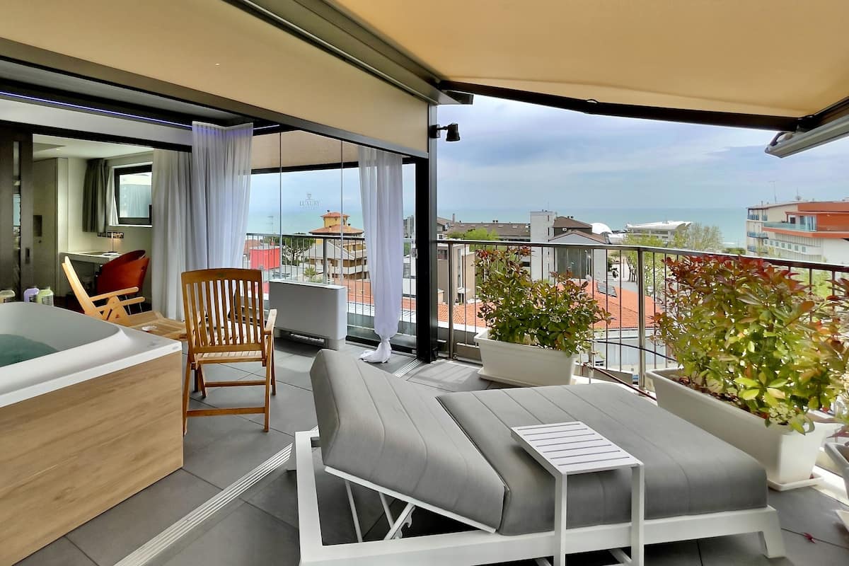 A balcony with a sofa, a chair, a small table, a potted plant, and a glass-enclosed area with a bathtub.
