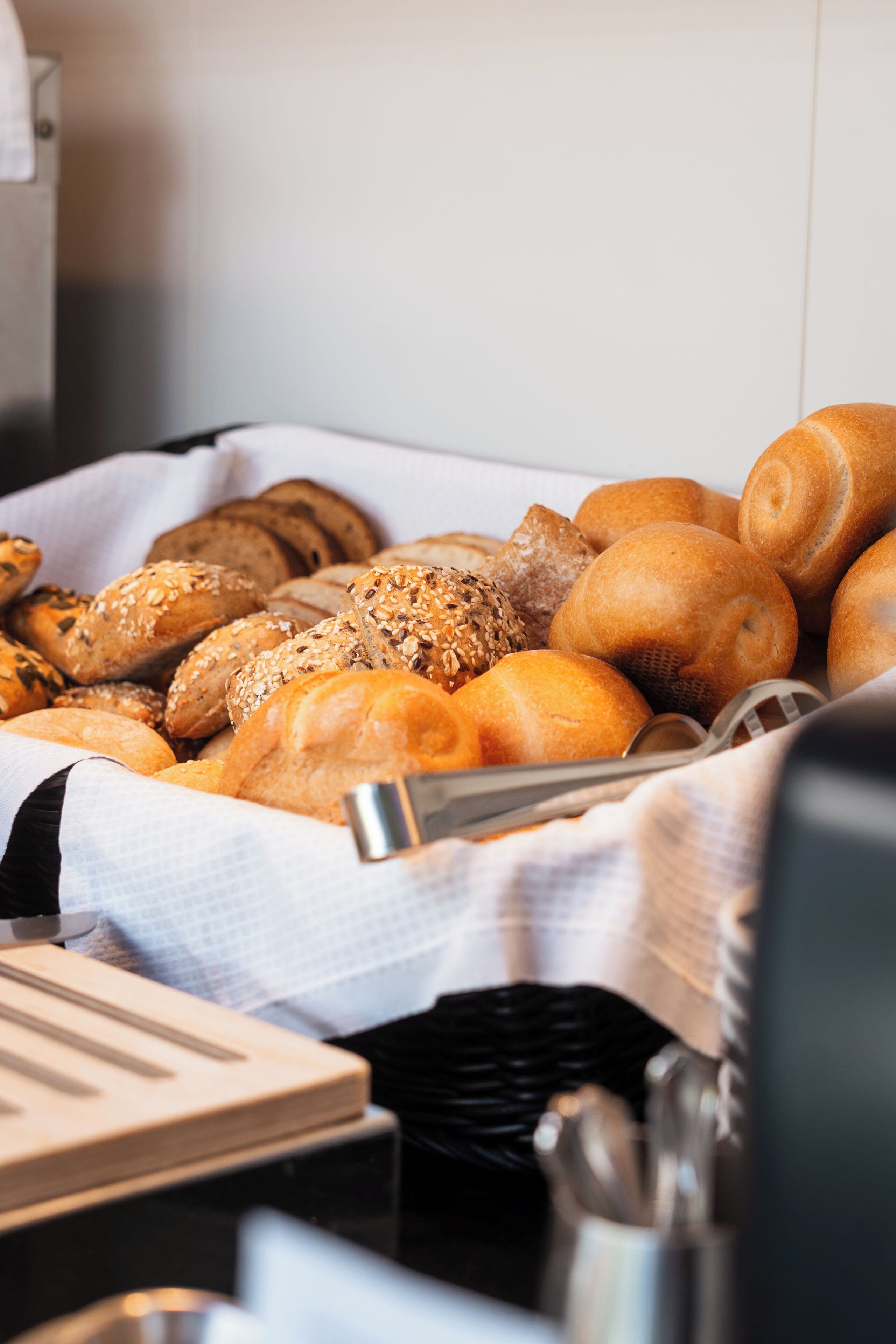 Eine Auswahl an Brotsorten und Brötchen in einem Korb, mit einem Küchenwerkzeug und einem Stoff in der Nähe.