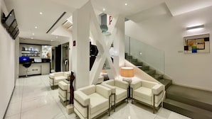 A modern living room with a sofa set, a glass staircase, and a kitchen in the background. - Monaco Suites Hotel (Lignano Sabbiadoro)
