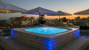 A hot tub with blue LED lighting, surrounded by wooden deck and lounge chairs, set against a cityscape at sunset. - Monaco Suites Hotel (Lignano Sabbiadoro)