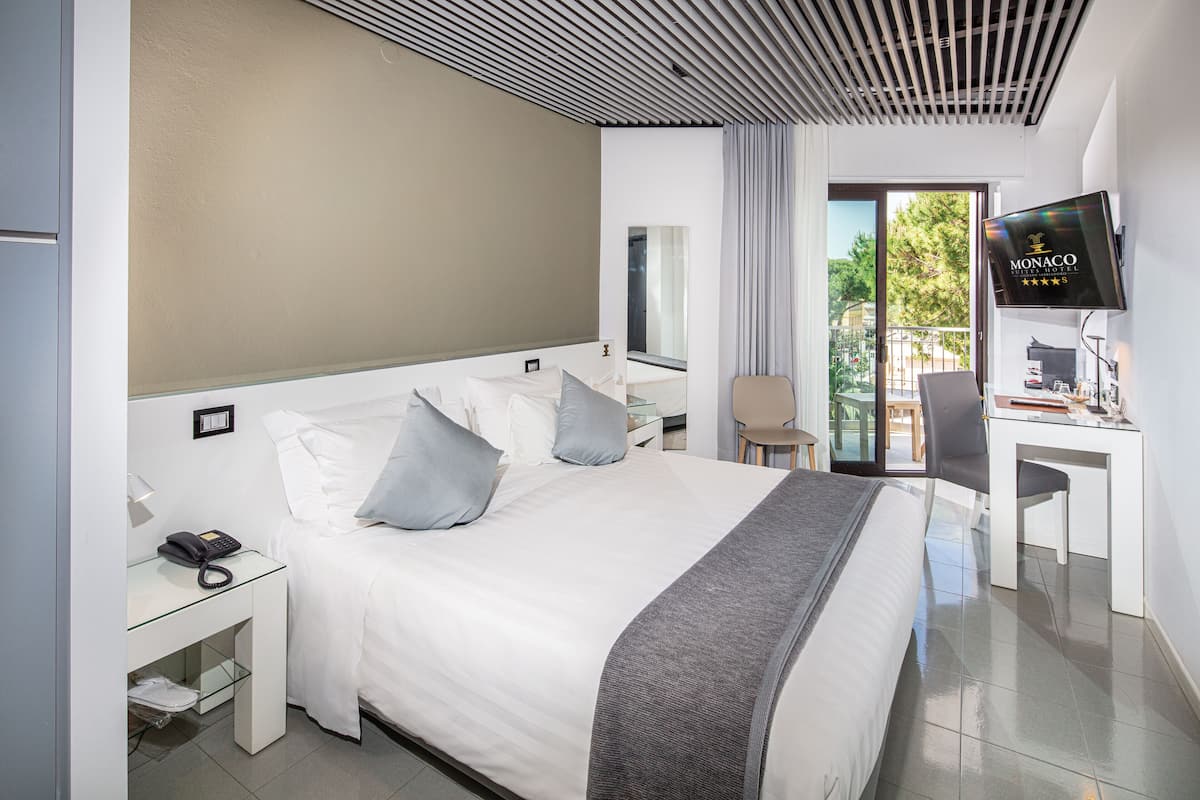 A modern hotel room with a large bed, a desk, a chair, a TV, and a balcony with a view of trees.