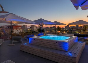 Terraza en la azotea con jacuzzi, mobiliario exterior, sombrillas y vistas a la ciudad al atardecer.