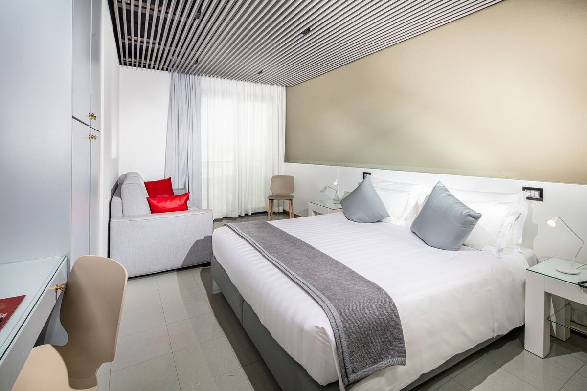 A modern hotel room with a large bed, a seating area with a red pillow, a desk with a lamp, and a large window with curtains.