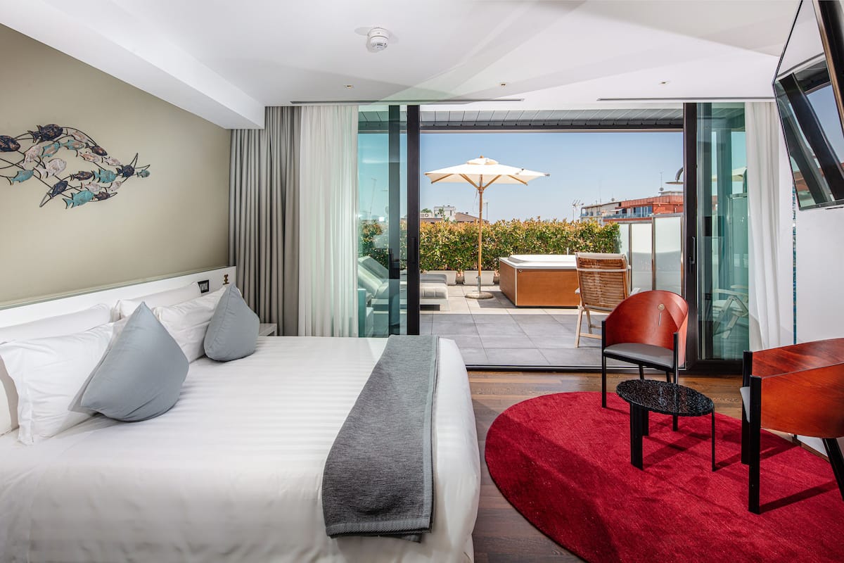 A modern hotel room with a large bed, a red rug, a dining area with a table and chairs, and a balcony with an umbrella.
