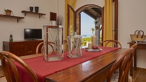 A dining area with a wooden table, red tablecloth, and chairs. There is a TV on a wooden cabinet, and a window with yellow curtains. - Villa Del Poggio (Castellammare del Golfo)