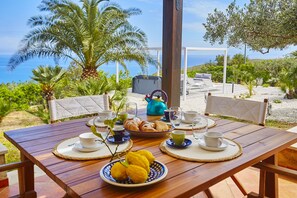 A wooden table set for breakfast with croissants, lemons, and cups, overlooking a scenic coastal view with palm trees and a white gazebo.