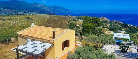 A yellow building with a tiled roof, a covered outdoor seating area, and a view of the sea and mountains.