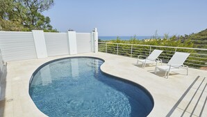 A swimming pool with a curved edge, two white lounge chairs, a white fence, and a view of the ocean. - Skrokkia (Alcamo)