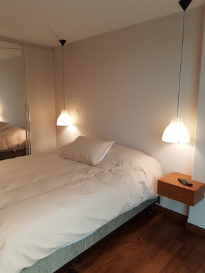 A neatly made bed with a pillow, a bedside table with a remote control, and a wall-mounted light fixture. - HB Calacoto (La Paz)