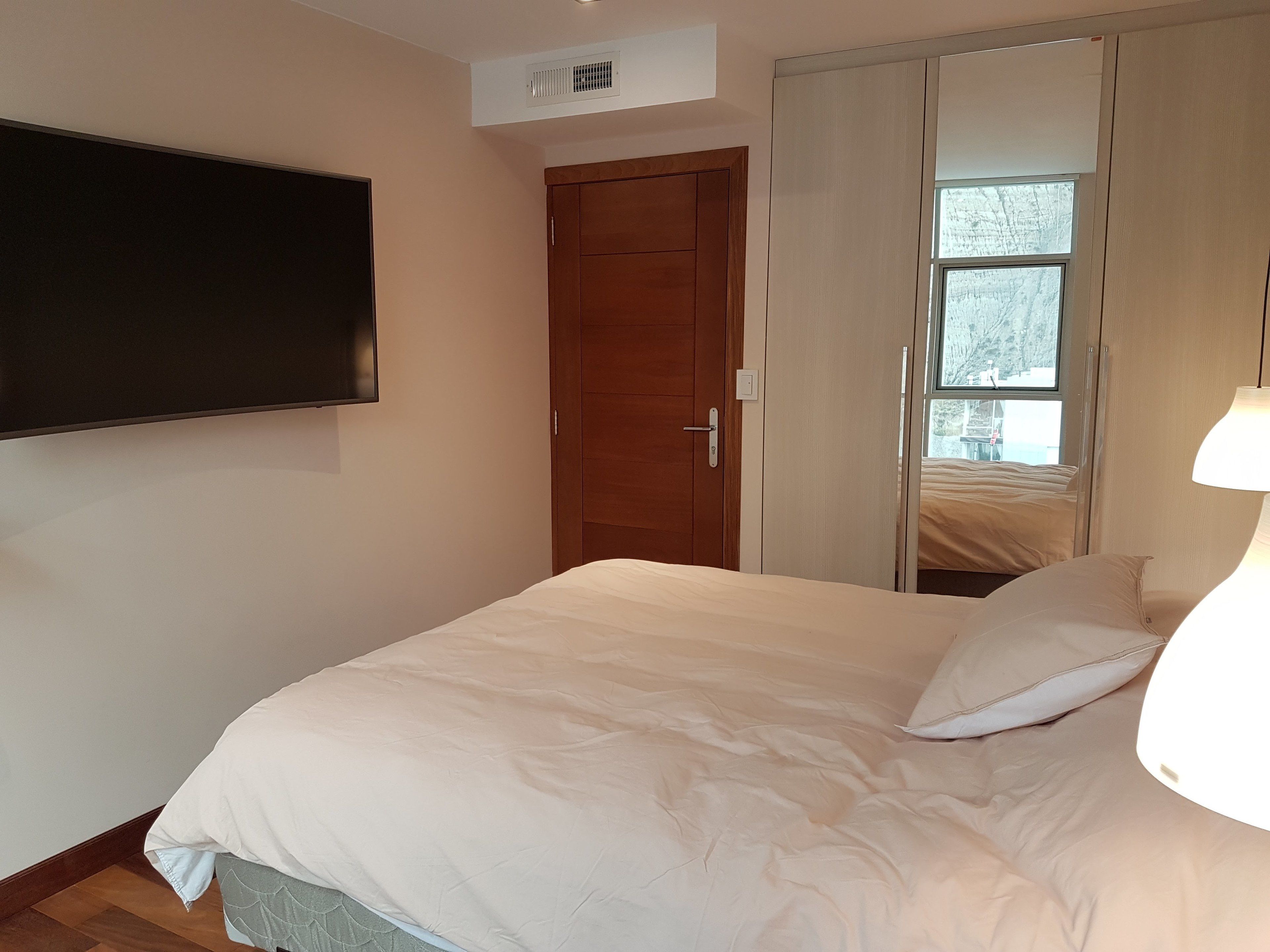 A bedroom with a bed, a TV mounted on the wall, a wardrobe, and a window.