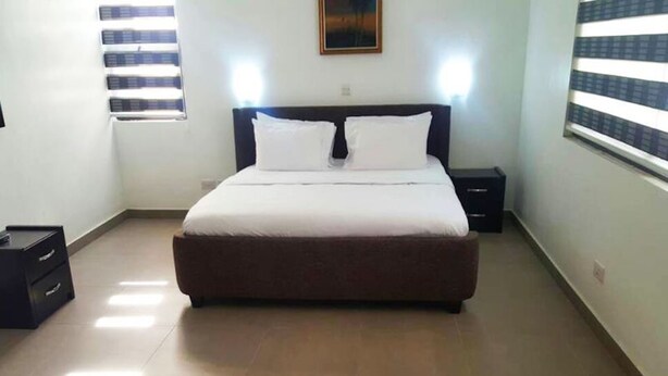 A bedroom with a bed, two bedside tables, a nightstand, a window with blinds, and a wall-mounted picture. - Gemini Place Apartments (Lagos)