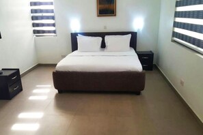 A bedroom with a bed, two bedside tables, a nightstand, a window with blinds, and a wall-mounted picture. - Gemini Place Apartments (Lagos)