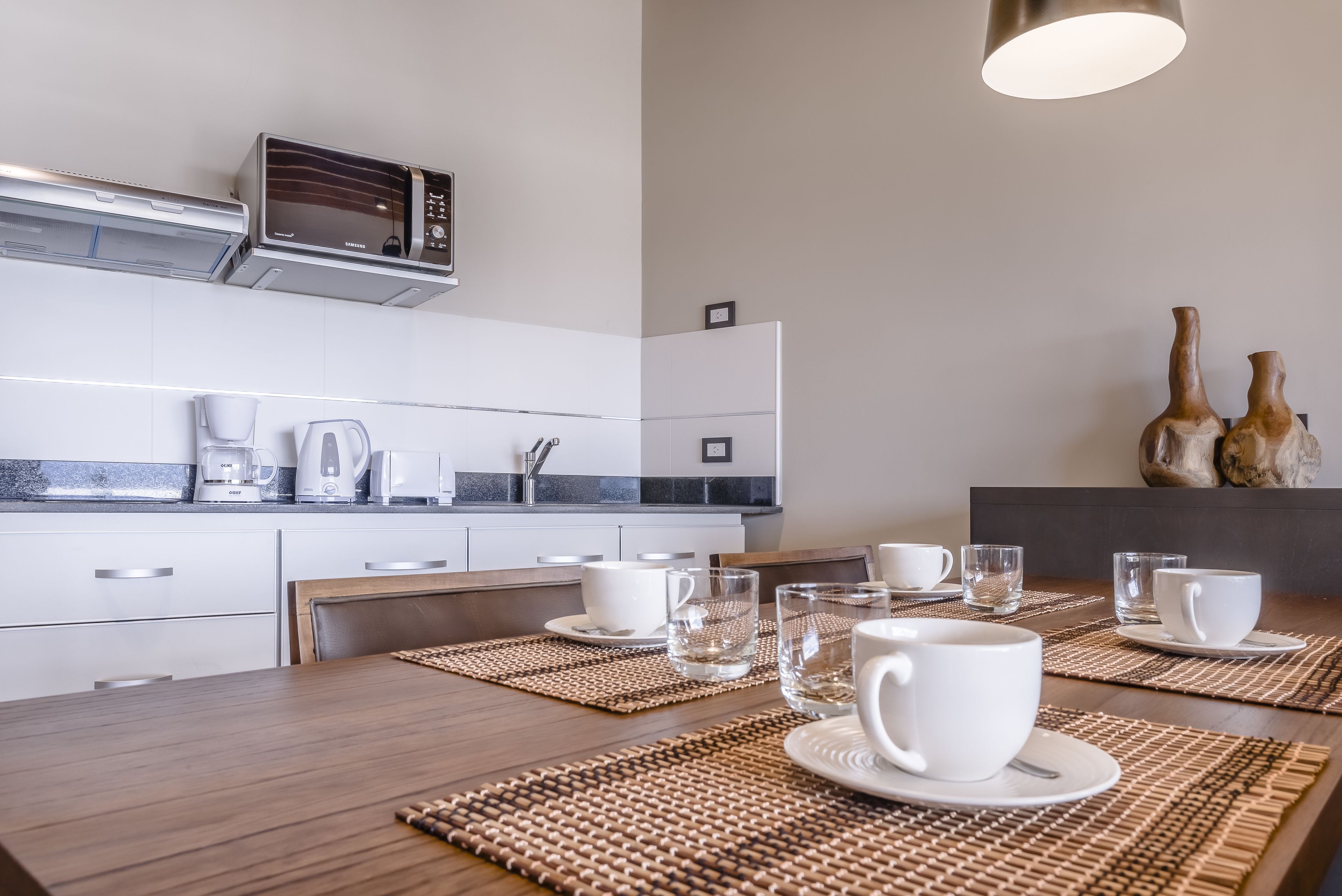 Una cocina moderna con microondas, cafetera y tostadora. Hay una mesa de comedor con manteles individuales y tazas blancas.