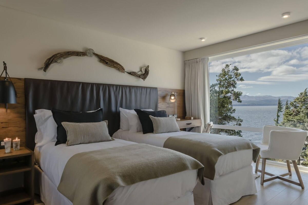 A bedroom with two beds, a nightstand, a lamp, a chair, and a view of the lake.