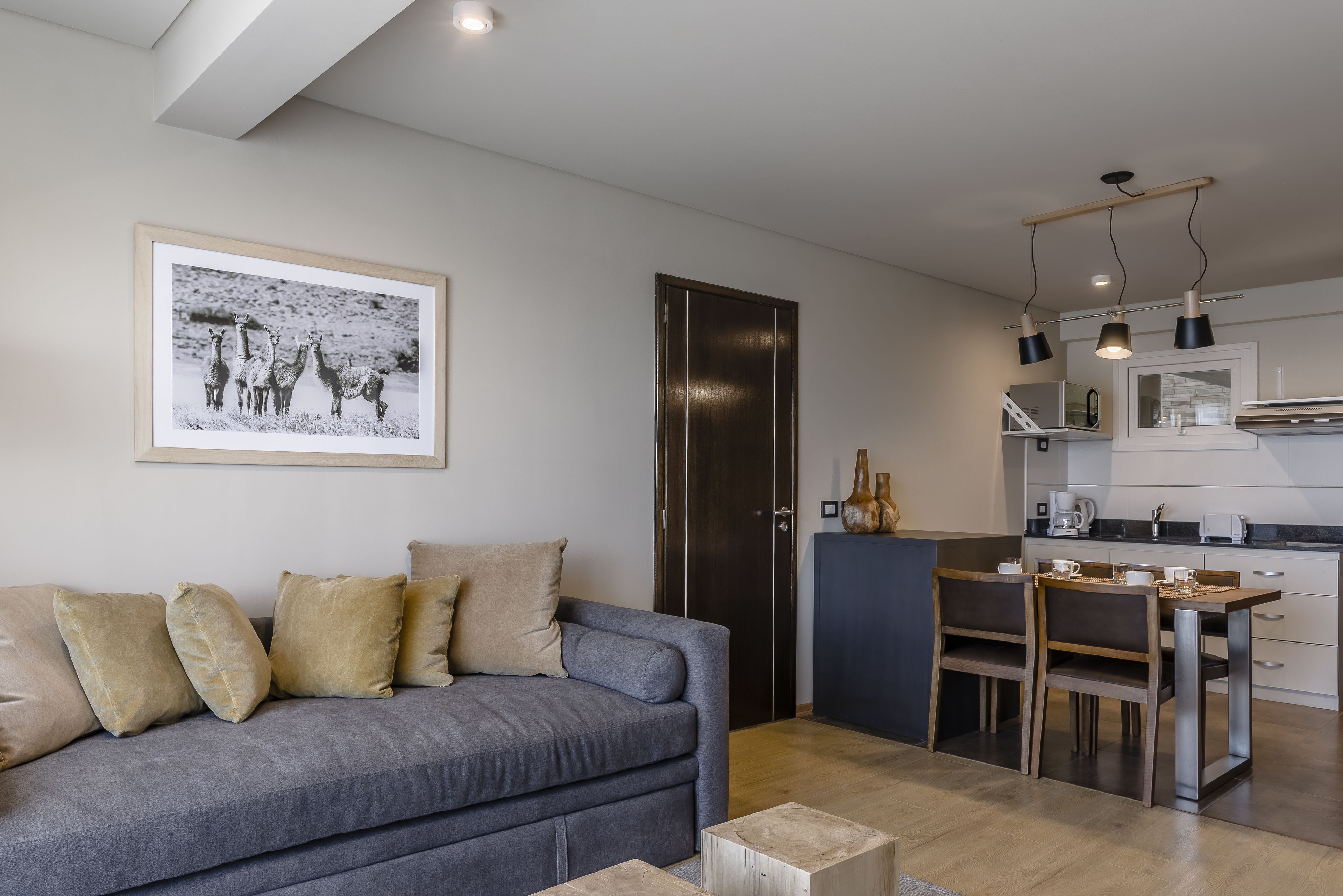 Una sala de estar moderna con un sofá gris, un comedor con mesa y sillas de madera, y una cocina con armarios blancos y encimera negra.