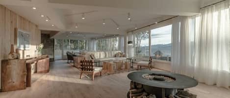 A spacious living room with a fireplace, wooden furniture, and large windows offering a view of the outdoors.
