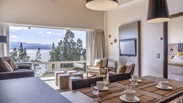 Una habitación moderna con un ventanal grande que da a un lago y montañas, amoblada con sofá, sillas y una mesa de comedor con tazas y platitos.