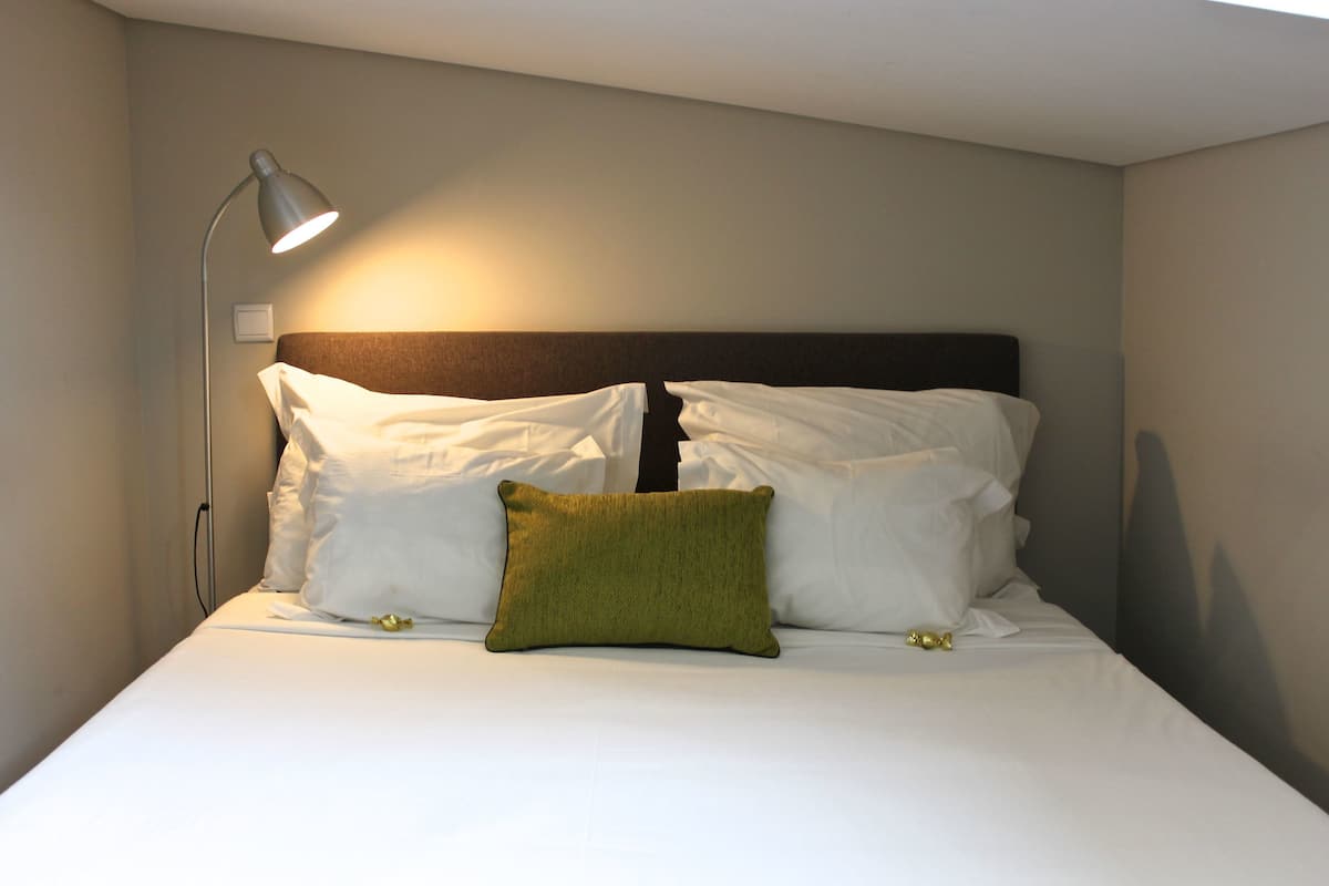 A neatly made bed with white and green pillows, a bedside lamp, and a plain wall.