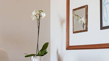 Un jarrón blanco con una orquídea sobre una mesa, un espejo enmarcado que refleja la orquídea y un texto enmarcado en la pared.