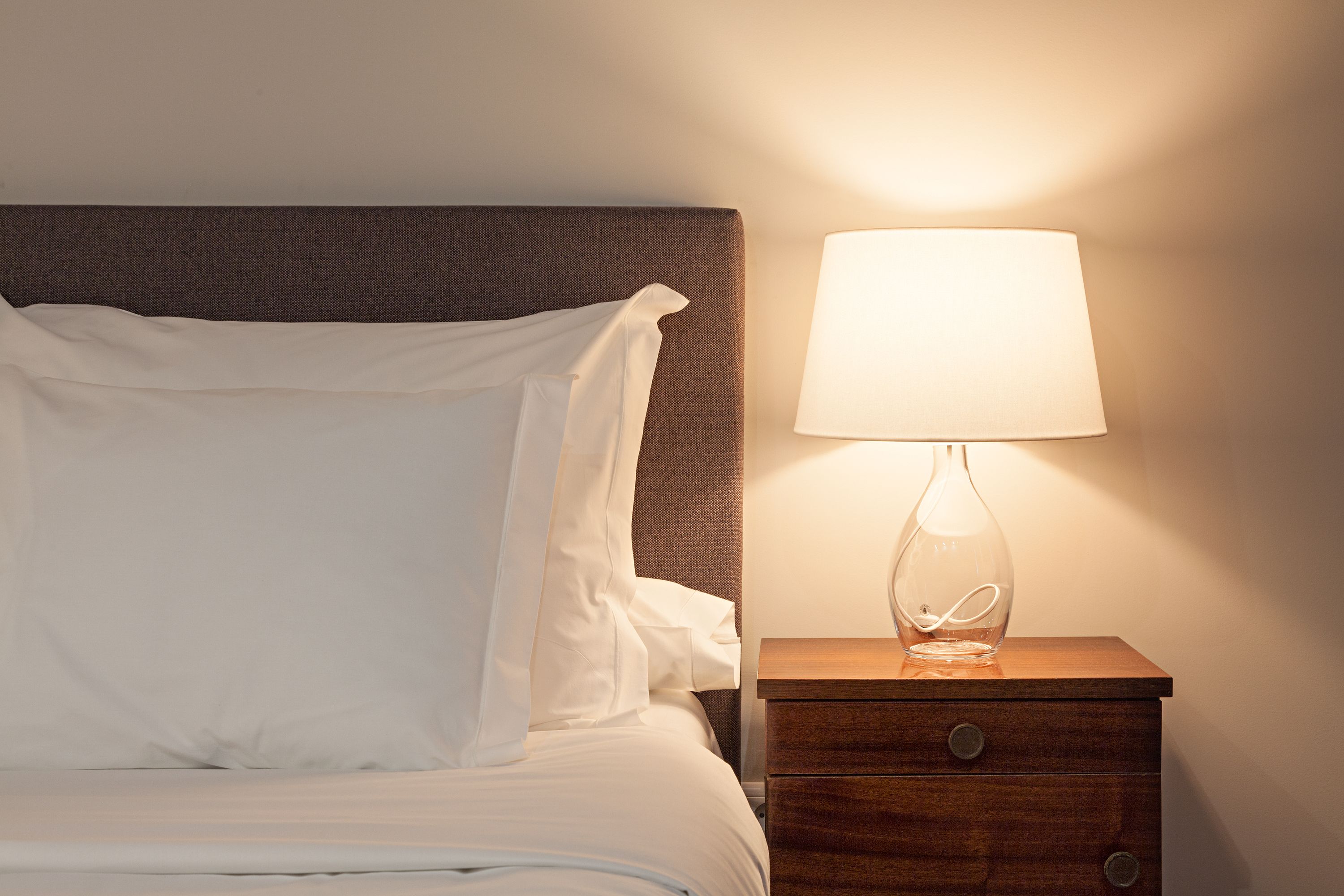A neatly made bed with white linens and pillows, a wooden bedside table with a lamp, and a drawer.