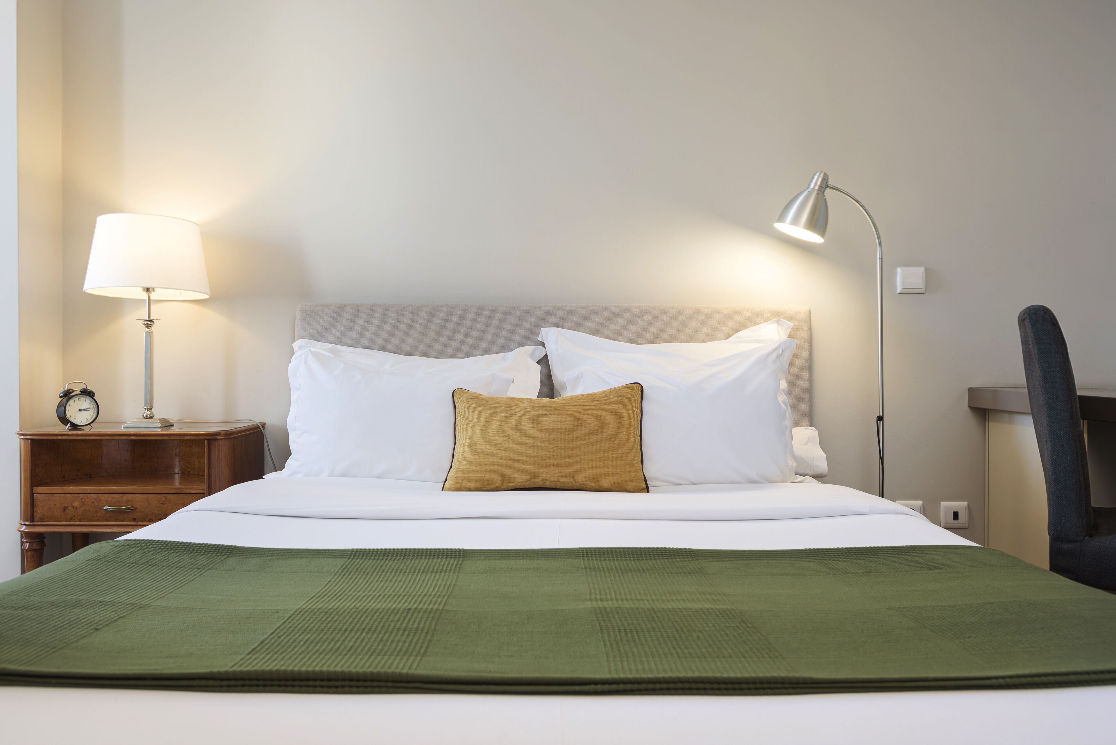 A neatly made bed with white linens and a green throw, a bedside lamp, and a nightstand with a clock.