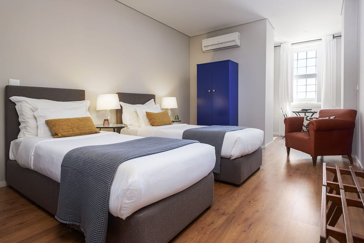 A hotel room with two beds, a blue wardrobe, a red armchair, and a wooden ladder.