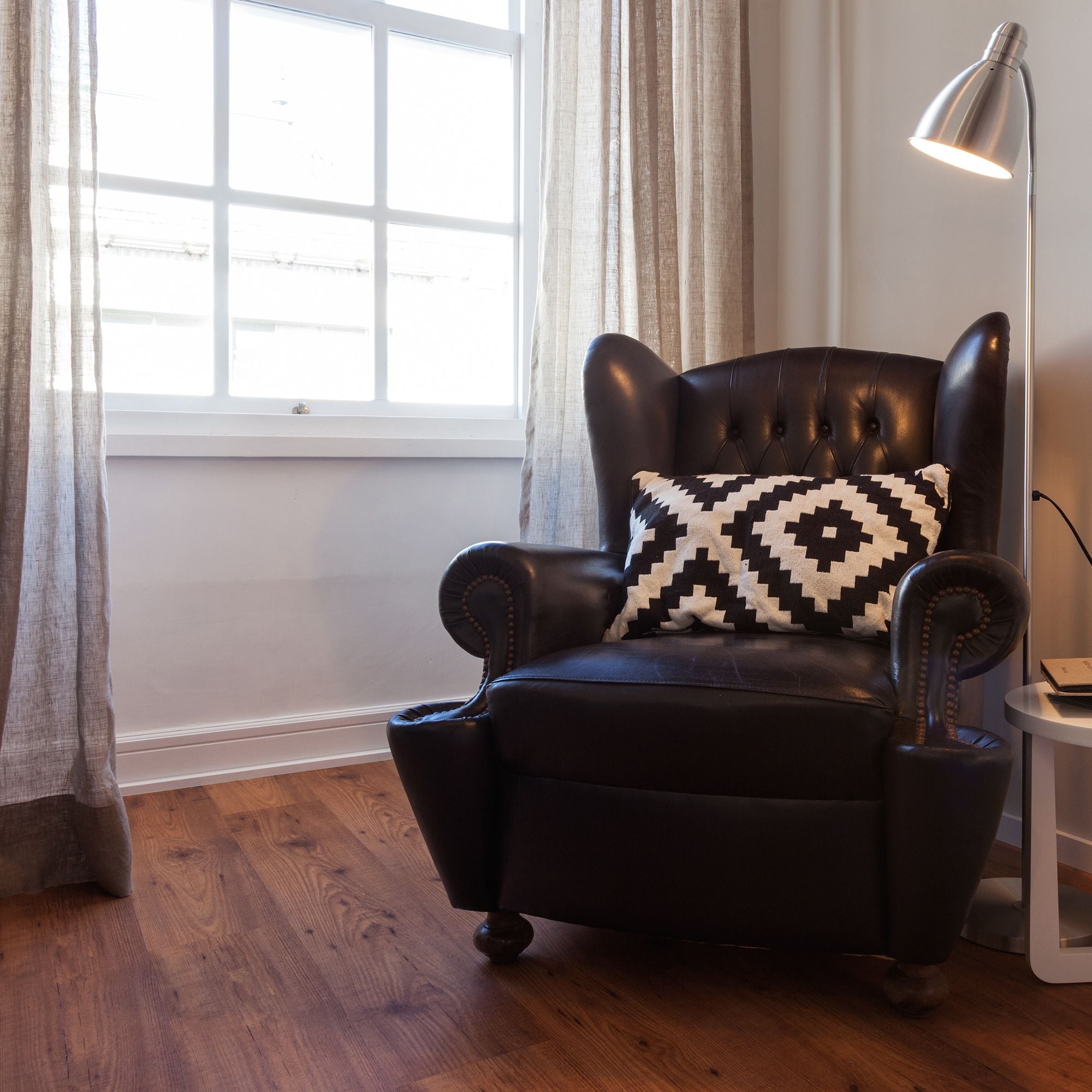 A black leather armchair with a patterned pillow, positioned next to a window with sheer curtains, a standing floor lamp, and a small side table.