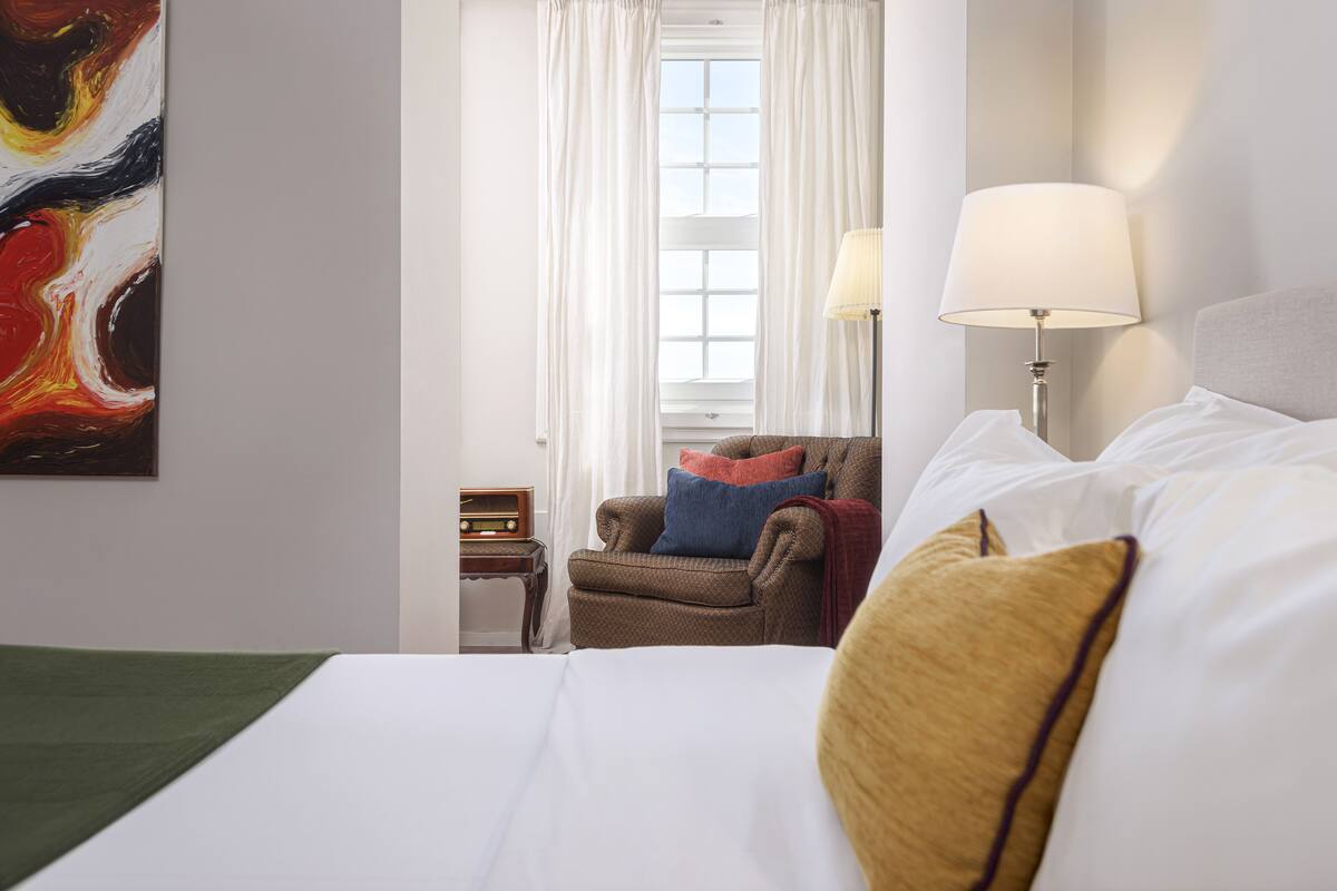 A hotel room with a bed, a chair, a lamp, a radio, and a window with curtains.