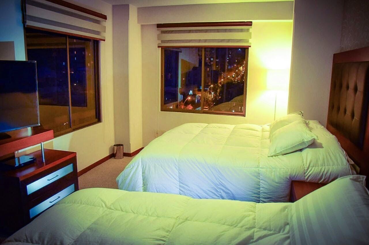 A hotel room with two beds, a TV on a wooden stand, and a window with a view of city lights.