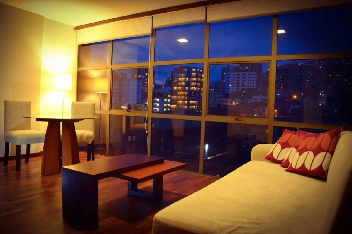 Una habitación de hotel moderna con un ventanal grande, un sofá, una mesa y dos sillas.