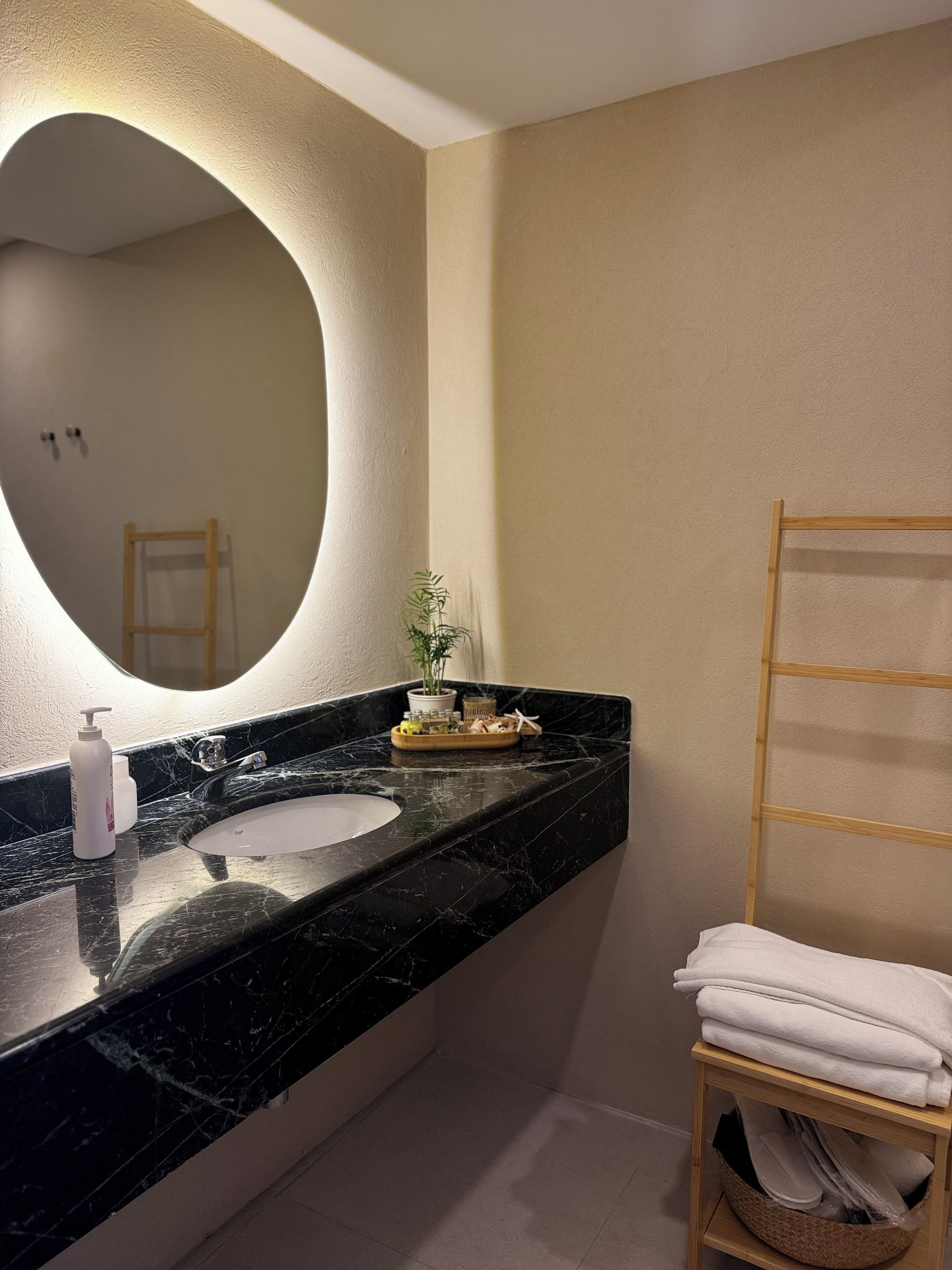 A bathroom with a black marble sink, a round mirror, a wooden towel rack, and a small potted plant.