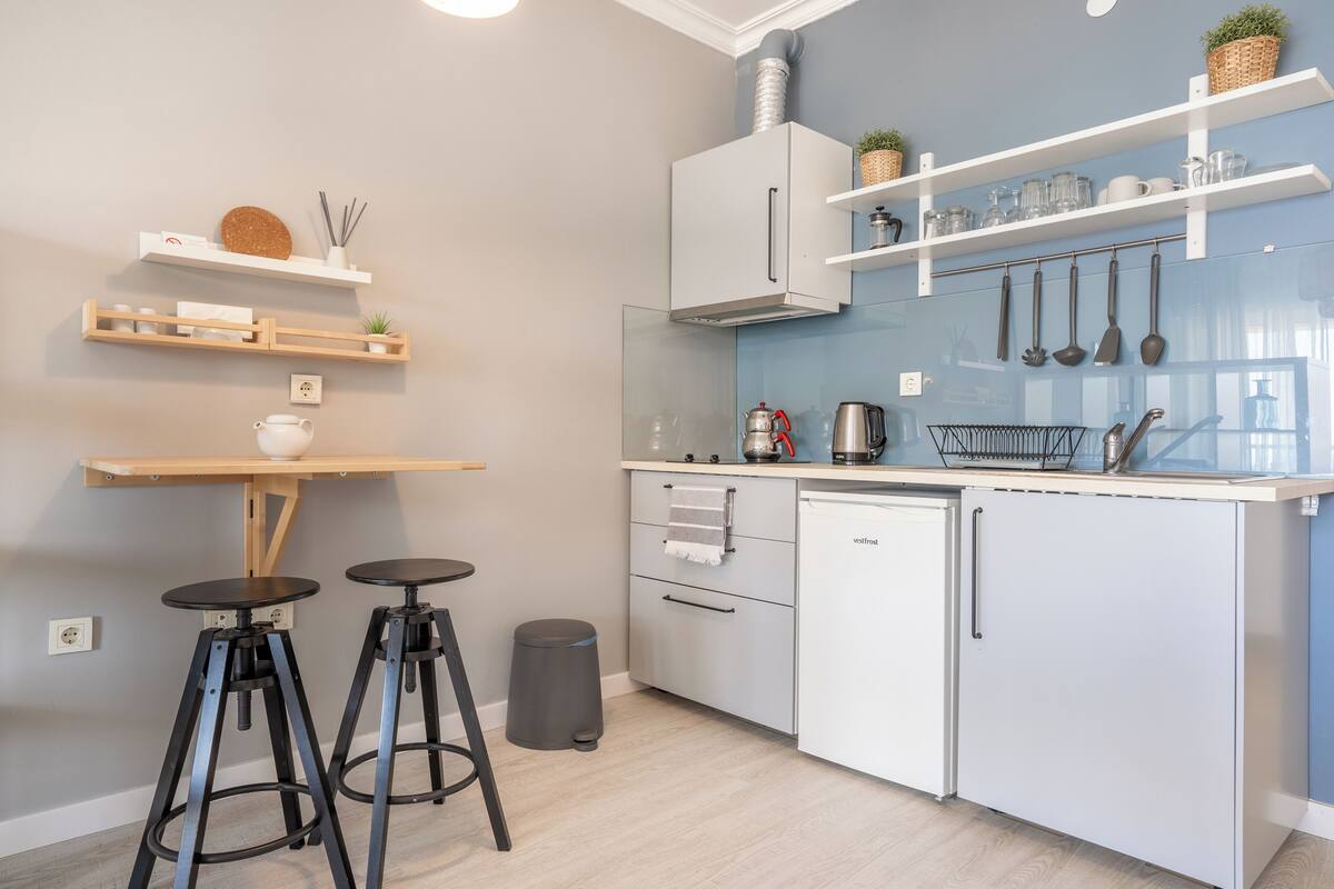 Una cocina moderna con armarios blancos, una estufa integrada y una cafetera. Hay dos taburetes y un cubo de basura.