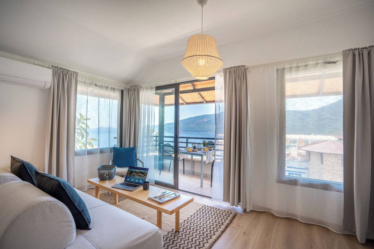 Una habitación de hotel moderna con un sofá, una mesa de centro con una computadora portátil y una revista, un ventanal con vistas al mar y un balcón con mobiliario exterior.