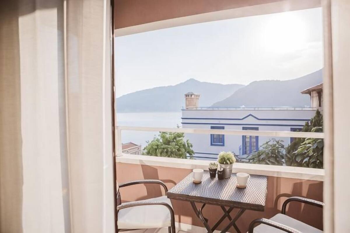 Un balcón con mesa y sillas, vista a un cuerpo de agua y montañas, y un edificio con fachada azul y blanca.