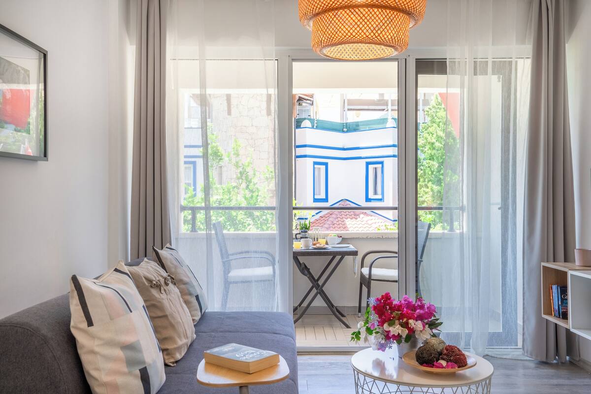 Una sala moderna con un sofá gris, una mesita redonda con un libro y flores, y un balcón con vistas a un edificio azul y vegetación.