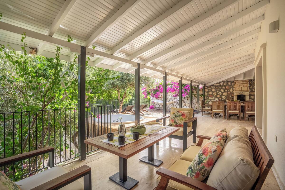 Un patio techado al aire libre con mesa de madera, asientos acolchados y un muro de piedra de fondo.