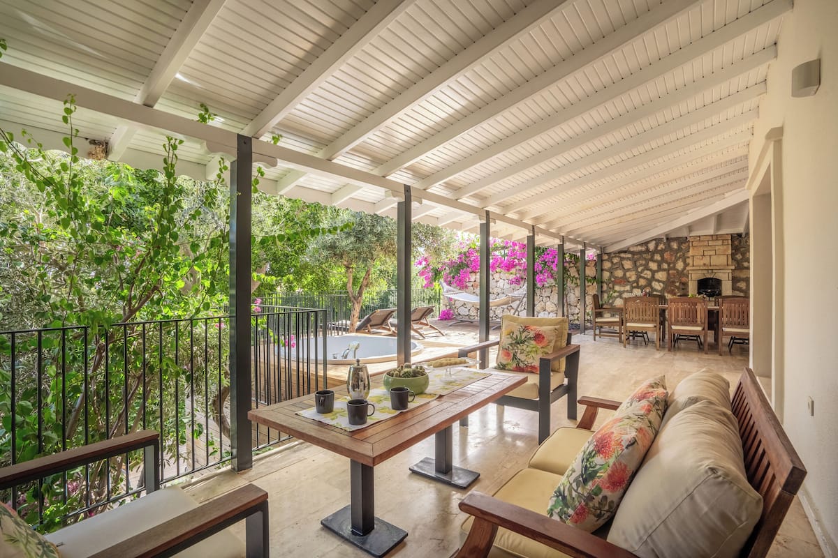 Un patio techado al aire libre con mesa de madera, asientos acolchados y un muro de piedra de fondo.