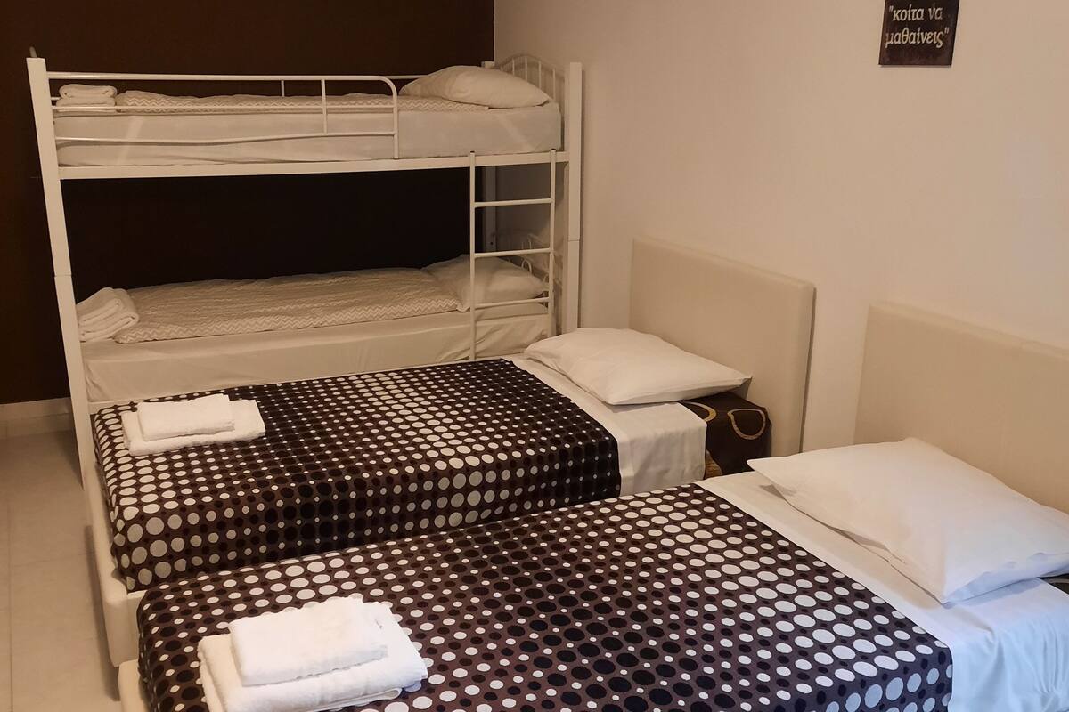 Habitación de hotel con dos camas, una con una colcha de lunares y la otra con una colcha blanca lisa. Hay una cama nido en la esquina y una mesita con una lámpara.