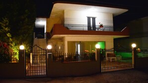 Una casa de dos pisos con un balcón, entrada protegida por un portón y un interior iluminado en verde.