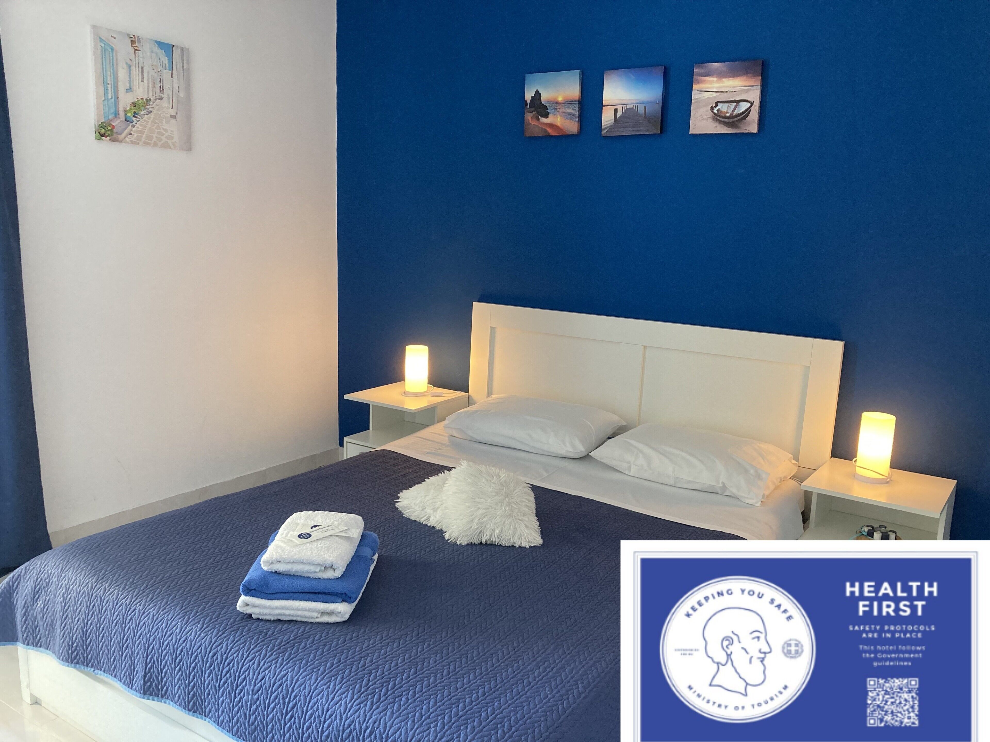 A hotel room with a blue wall, white bed, and artwork. There are towels on the bed and a sign about health protocols.